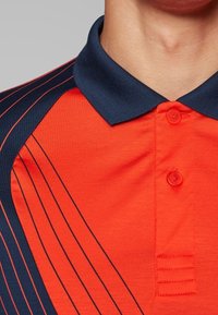 Červené polo tričko s námořnickými akcenty, s texturovaným límcem, knoflíkovou légou a diagonálními pruhy v kontrastní námořnické a oranžové barvě.