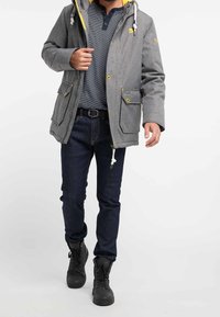 Uomo che indossa una giacca a hood grigia, una camicia a righe blu navy, jeans scuri, una cintura nera e scarpe alte nere che cammina su uno sfondo bianco.