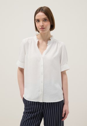 Donna con capelli castani corti che indossa una camicetta bianca a maniche corte con bottoni e pantaloni blu navy a righe sottili, in piedi davanti a uno sfondo semplice.