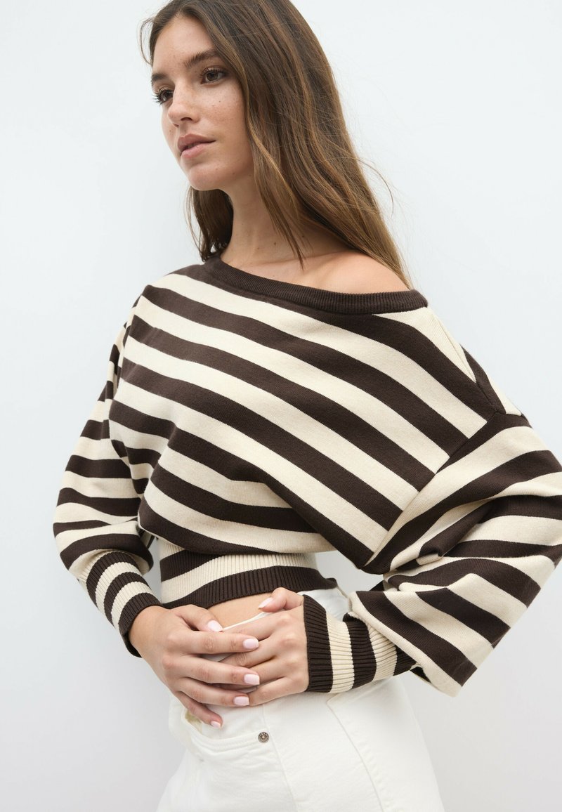 Young woman with long brown hair wears an off-shoulder brown and white striped sweater and white pants, posing with hands clasped at waist.