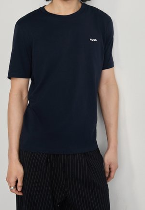 Person, die ein dunkelblaues Hugo-T-Shirt und schwarze Nadelstreifenhose trägt, steht vor einem schlichten hellen Hintergrund.