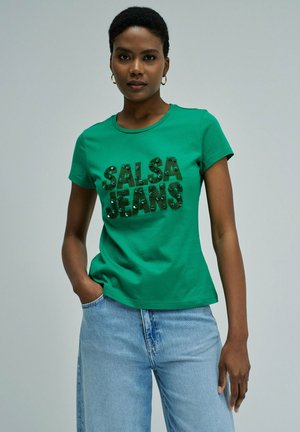 Femme aux cheveux courts portant un t-shirt vert "SALSA JEANS" et un jean bleu clair, debout avec une main dans la poche devant un fond uni.