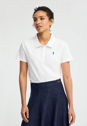 Kobieta ubrana w białą koszulkę polo z krótkim rękawem z małym czarnym logo i ciemnoniebieską spódnicę z wysokim stanem, stojąca na jednolitym tle.