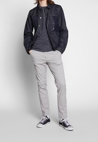 Jeansjacka i mörkblått med en ficka i patchstil, lager på lager över en mörkgrå prickig skjorta. Ljusgrå slim-fit byxor och svarta sneakers.