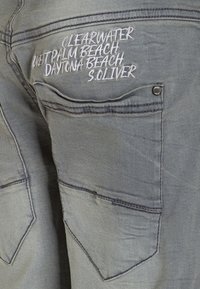 Šedé denimové kraťasy s dvěma zadními kapsami, z nichž jedna má bílé vyšívané texty, a malou černou kovovou knoflíkovou ozdobu.