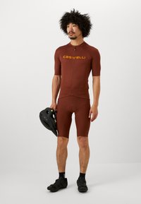 Braunes Radtrikot mit kurzen Ärmeln und Shorts, mit einem Reißverschluss vorne und dem "Castelli"-Logo in Gelb. Schwarzer Helm wird in einer Hand gehalten.