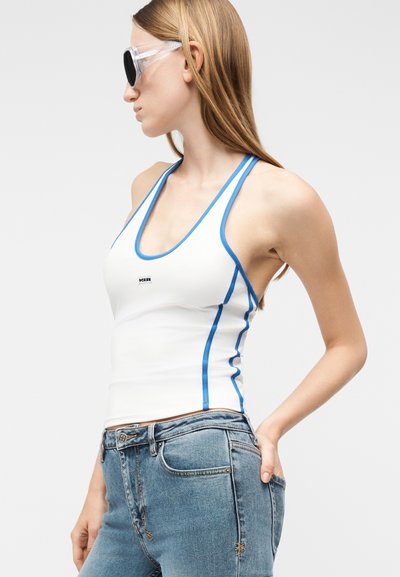 Νέα γυναίκα που φοράει λευκή αμάνικη μπλούζα με μπλε περιθώριο, μπλε τζιν και διαφανή γυαλιά ηλίου, στέκεται σε πλευρικό προφίλ.