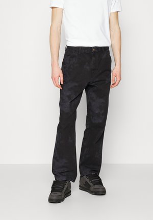 Man draagt zwarte tie-dye rechte broek, zwarte sneakers en een wit T-shirt met korte mouwen, staand tegen een effen achtergrond.