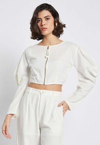 Weißes kurzes Blusenshirt mit langen Puffärmeln, Rundhalsausschnitt und Frontreißverschluss. Hergestellt aus einem strukturierten Stoff mit glatter Oberfläche.