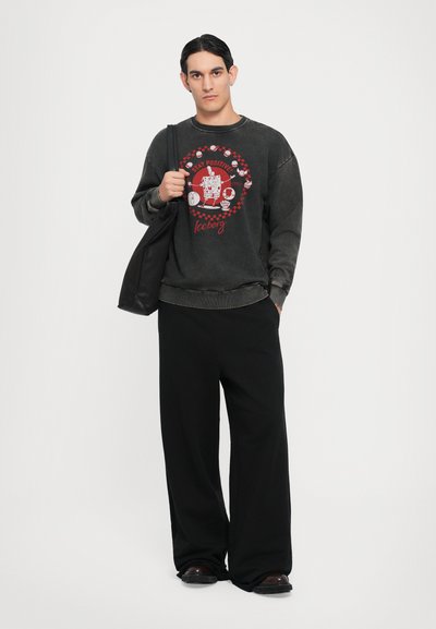 Schwarzer Sweatshirt mit Rundhalsausschnitt und rotem grafischen Design, kombiniert mit lockeren schwarzen Hosen. Modell hält eine schwarze Tragetasche.