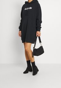 Robe sweat à capuche noire avec logo blanc, manches longues et capuche à cordon. Associée à des bottines noires et un petit sac à main noir.