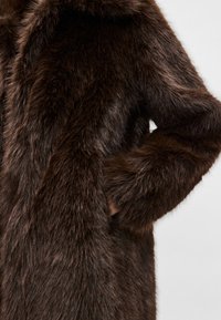 Hnedý kabát z umelej kožušiny s jemnou, hustou textúrou, vybavený kapucňou a bočným vreckom. Kožušina má prirodzený lesk, ktorý zvyšuje jej vizuálnu hĺbku.