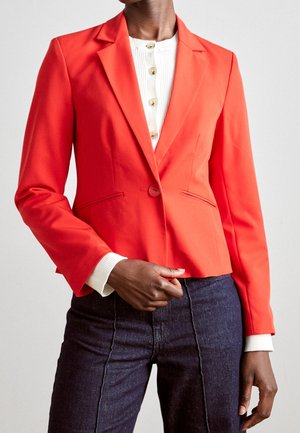 Vrouw die een aansluitende felrode blazer draagt over een wit knooptopje en donkere spijkerbroek, staand tegen een effen achtergrond.