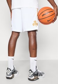 Baltas basketbola šorts ar zelta āboliņa logotipu. Melni un balti sporta apavi ar lauvas grafiku uz papēža. Oranžs basketbols rokā.