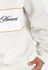 Grijze sweatshirt met witte en gele accenten, met "Moment" in zwarte borduring. Zachte stof met ribbelmanchetten en onderrand.