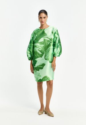 Woman wearing green floral knee-length dress with puffed sleeves and leopard print low-heeled shoes, standing against white background.