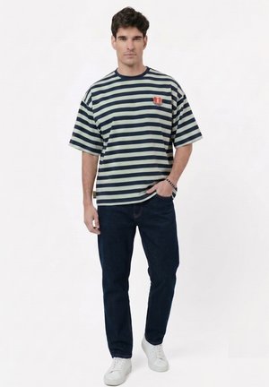 Hombre con camiseta de rayas azul marino y gris claro, vaqueros oscuros, zapatillas blancas, de pie con la mano parcialmente en el bolsillo frente a fondo blanco.