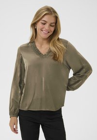Olijfgroene blouse van glad, glanzend materiaal met lange mouwen en een v-hals, met een rimpelende halslijn. Gekoeld met zwarte broek.