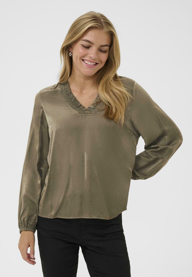 Olijfgroene blouse van glad, glanzend materiaal met lange mouwen en een v-hals, met een rimpelende halslijn. Gekoeld met zwarte broek.