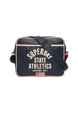 Sac d'épaule rectangulaire bleu marine avec bordure crème, bandoulière réglable, texte "SUPERDRY STATE ATHLETICS COACHING DEPT." et petit graphique du drapeau américain.