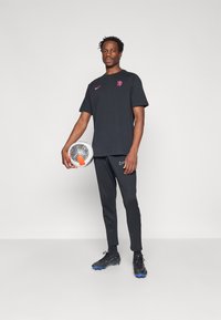 Tricou Nike negru cu logo roz, pantaloni negri asortați și ghete negre cu accente albastre. Ține în mână o minge de fotbal cu un model geometric.