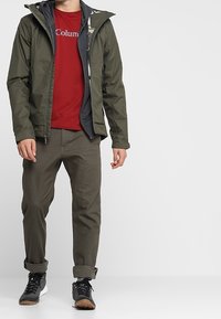 Herreudendørsjakke i mørk olivengrøn med hætte, lagdelt over en rød skjorte. Kombineret med olivengrønne bukser og sorte sko.