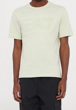 Man wearing light beige short-sleeve t-shirt with embossed "EA7" logo and black cargo-style pants against plain background.