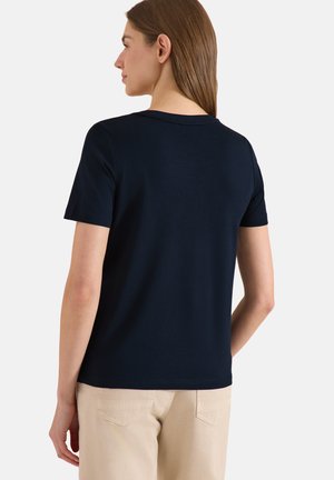 Donna con capelli castani lisci che indossa una maglietta blu scuro a maniche corte e pantaloni beige, di spalle su uno sfondo chiaro e semplice.