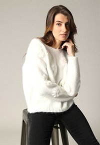 Witte, donzige gebreide trui met getextureerde bloemen details op de mouwen, ronde halslijn en geribde boorden, gecombineerd met zwarte skinny jeans.