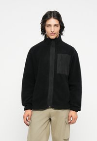Schwarze Fleece-Jacke mit hohem Kragen und Frontreißverschluss. Sie hat eine strukturierte Oberfläche und eine kontrastierende schwarze Tasche auf der Brust.
