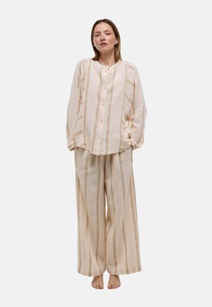Femme debout pieds nus portant une chemise beige rayée ample boutonnée et un pantalon large assorti sur fond blanc.