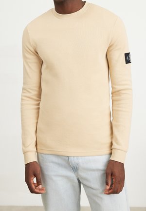 Beige gestricktes Langarmshirt mit Rundhalsausschnitt und einem schwarzen Logo-Patch auf dem linken Ärmel. Glatte Textur mit schmaler Passform.