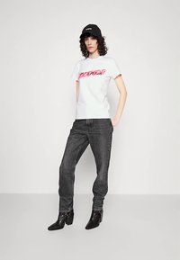 Ung person iført en hvid t-shirt med grafik, mørkegrå jeans, sort kasket og sorte ankelstøvler med hæl, stående foran en ensfarvet hvid baggrund.