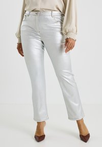 Jean métallisé argenté à coupe slim, avec une taille mi-haute, des poches avant et des coutures ton sur ton. Porté avec un chemisier crème et des talons marron.