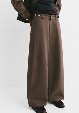 Osoba nosząca luźne, szerokie brązowe spodnie z przodu zapinane na guziki i czarne buty, stojąca na tle jednolitego jasnego tła.