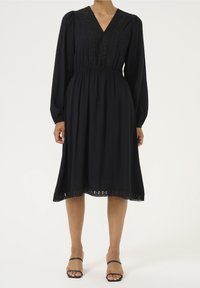 Robe noire à manches longues, col en V et détails en dentelle le long du corsage et de l'ourlet. Tissu lisse, longueur genou, avec une coupe décontractée.