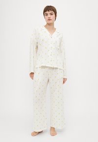 White pajama set with yellow lemon prints, featuring a button-up shirt and loose-fitting pants. Textured fabric with a relaxed fit.