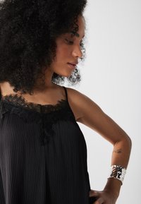 Camisola nera con bordo in pizzo e pieghe verticali, dotata di spalline sottili. Modella che indossa un braccialetto in metallo argentato al polso.