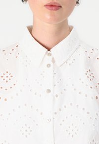 Witte katoenen shirt met ingewikkelde borduursels en uitsnijdingen, met een button-down kraag en knopen aan de voorkant. Glad, lichtgewicht textuur.