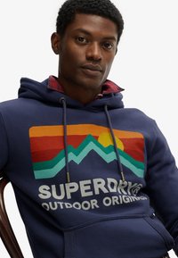Marine hoodie met grafische print waarop bergen, een zon en de tekst "SUPERDRY OUTDOOR ORIGINALS" staan. Met trekkoorden en detail van een voorzak.
