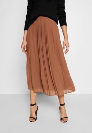 Mujer con falda midi plisada marrón, blusa negra de manga larga y tacones altos con tiras en el tobillo, sobre un fondo liso.