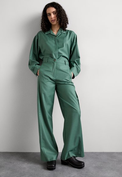 Completo due pezzi in satin verde composto da una camicia con colletto e bottoni e pantaloni larghi a vita alta. Le zoccoli neri completano il look.