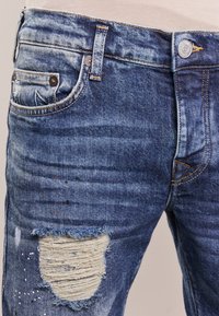 Siniset kuluneet farkut, joissa on haalistunut pesu, kuluneet alueet ja iso revitty polvi. Ominaisuuksiin kuuluvat klassinen viiden taskun tyyli ja metallipainike.