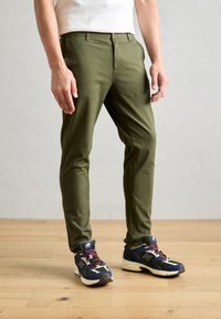 Olijfgroene slim-fit broek met een vlakke voorkant, ritssluiting, zijzakken, gedragen met donkere sneakers met meerkleurige accenten.