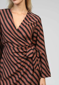 Femme portant une robe enroulée à rayures diagonales marron et noire, avec un nœud à la taille, des manches longues et un décolleté en V.