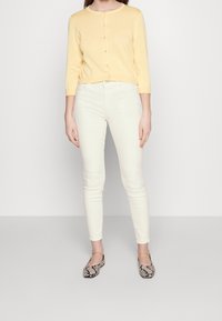 Gul, langærmet cardigan med rund halsudskæring og knapper, parret med tætsiddende hvide jeans og flade sko i sort og hvid slangeskin.