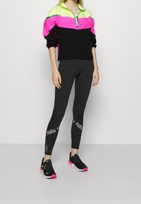 Colorful athletic jacket in neon pink, yellow, and black, paired with fitted black leggings featuring graphic accents and black sneakers with pink details.