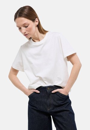 Femme aux cheveux bruns raides portant un t-shirt blanc uni à manches courtes et un jean taille haute bleu foncé, les mains dans les poches, regardant vers le bas.