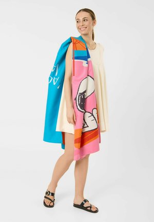 Frau in einem ärmellosen cremefarbenen Kleid und schwarzen Sandalen, die einen bunten Schal mit blauem, pinkem und orangefarbenem Muster über die Schulter legt, lächelt und schaut nach rechts.