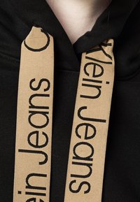 Czarna bluza z kapturem z beżowymi sznurkami, na których widnieje napis „Klein Jeans” oraz „super” wytłuszczonymi czarnymi literami. Miękki materiał, casualowy styl.